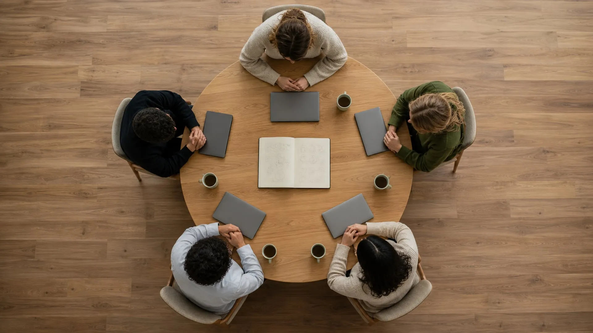 Overhead view of five people seated evenly around a round table, each the same distance from a single open notebook at the center, all working from one shared source.